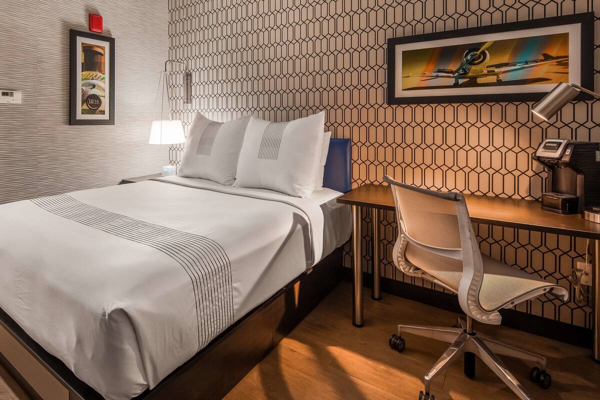 A hotel room with a bed, desk, chair, and a wall with a geometric pattern.