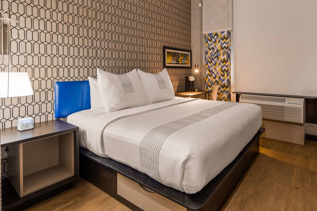 A modern hotel room with a neatly made bed, a bedside table with a lamp, a desk, and a chair.