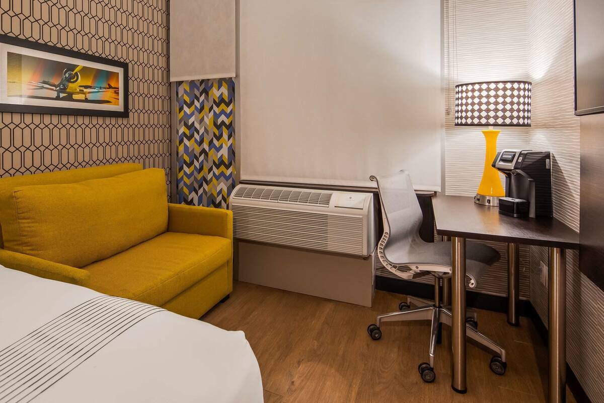 A hotel room with a bed, a yellow sofa, a desk with a chair, a coffee machine, and a wall-mounted air conditioner.