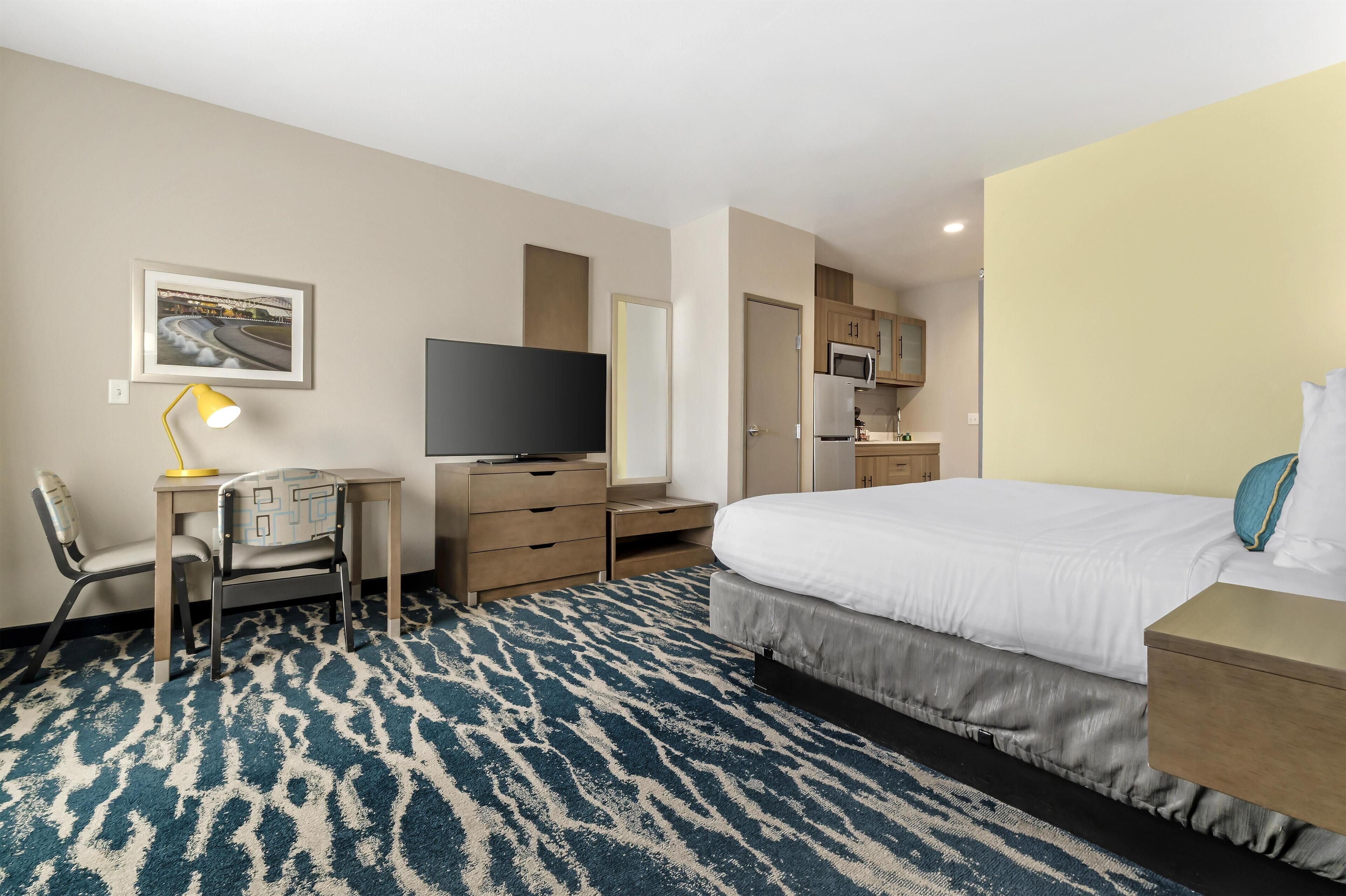 A hotel room with a bed, a desk with a chair, a television, a microwave, and a small kitchenette.