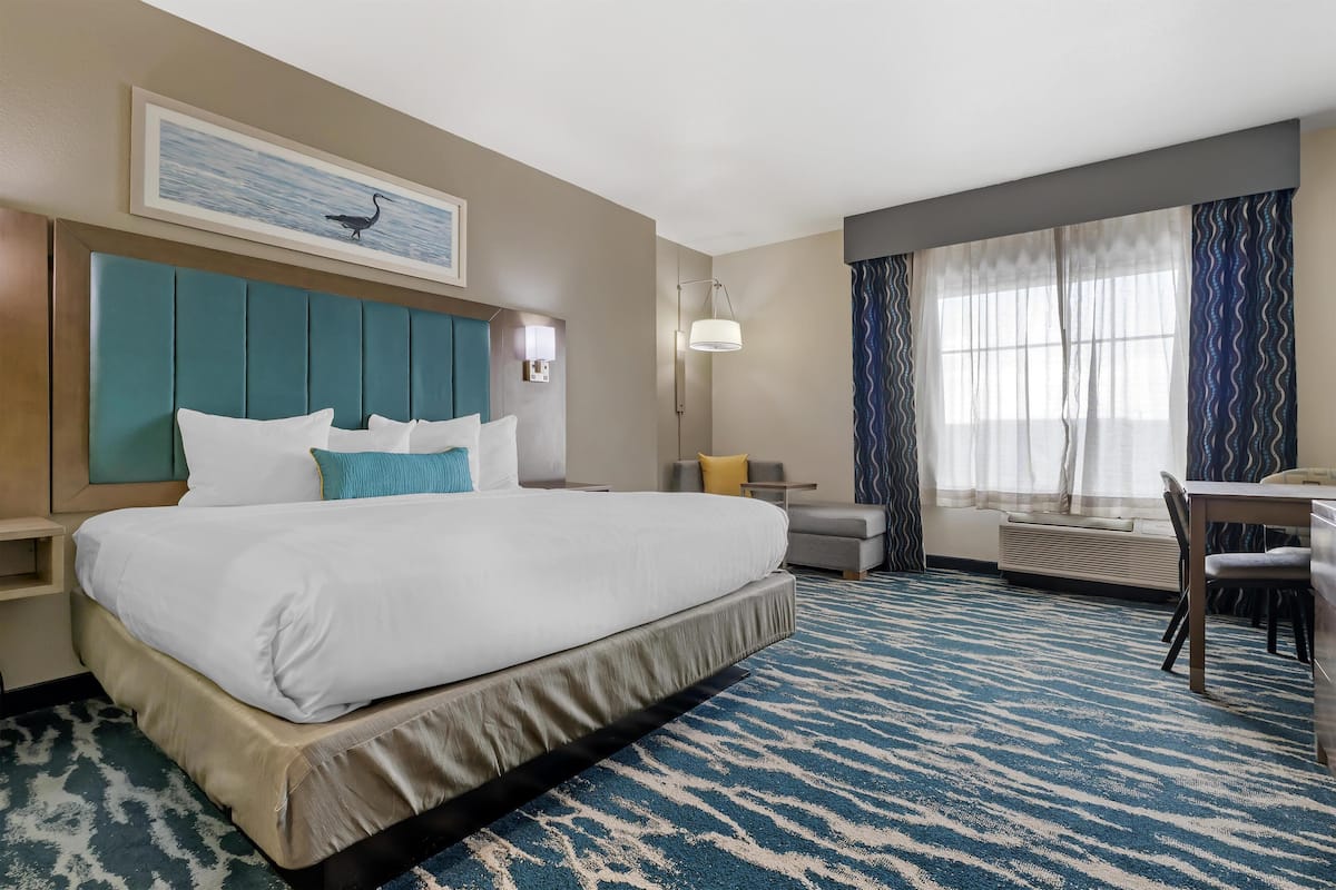 Habitación de hotel con una cama grande, un escritorio, una silla, una ventana con cortinas, una lámpara de pared y un cuadro enmarcado sobre la cama.