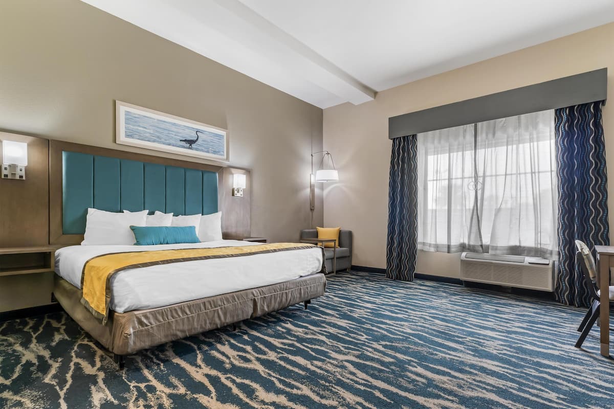 Habitación de hotel con una cama grande, cabecero, mesita de noche, lámpara, ventana con cortinas y una silla.