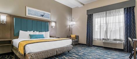 A hotel room with a large bed, a headboard, a bedside table, a lamp, a window with curtains, and a chair.