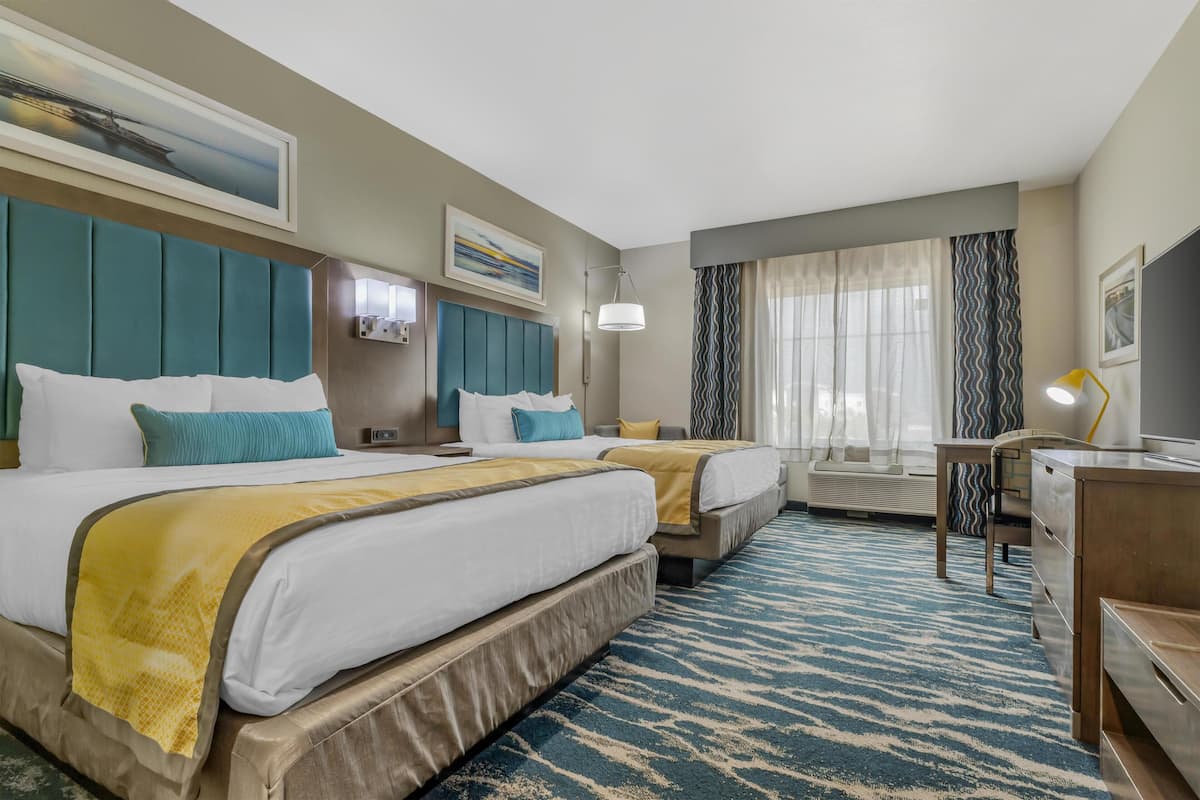 Habitación de hotel con dos camas, un escritorio, una silla, una lámpara y un ventanal grande con cortinas.