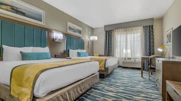Habitación de hotel con dos camas, un escritorio, una silla, una lámpara y un ventanal grande con cortinas.