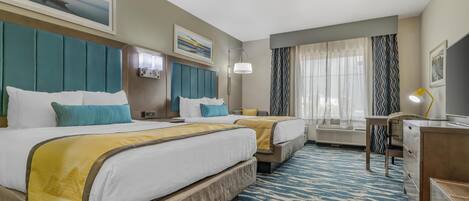 Habitación de hotel con dos camas, un escritorio, una silla, una lámpara y un ventanal grande con cortinas.