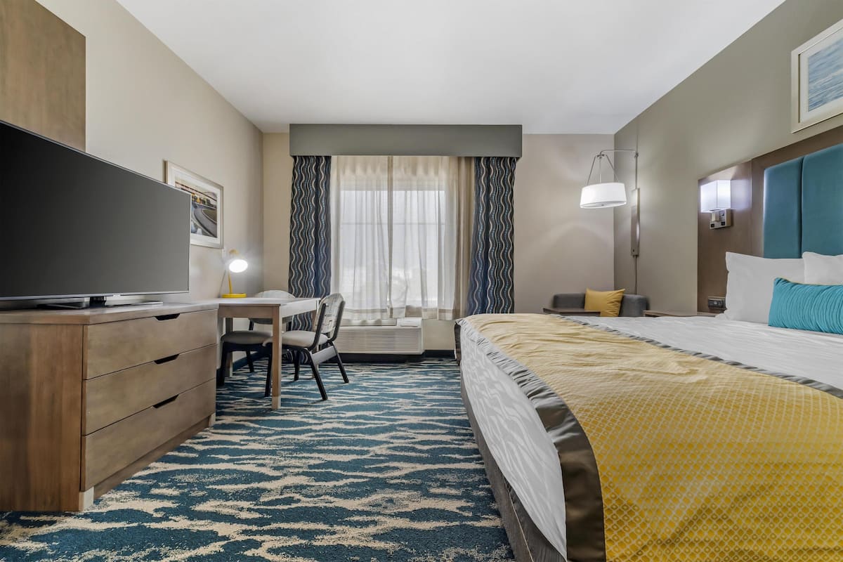 Una habitación de hotel con cama, escritorio, silla, televisión y ventana con cortinas.