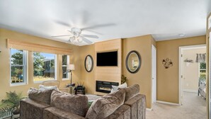 A living room with a fireplace, a ceiling fan, and a large window.