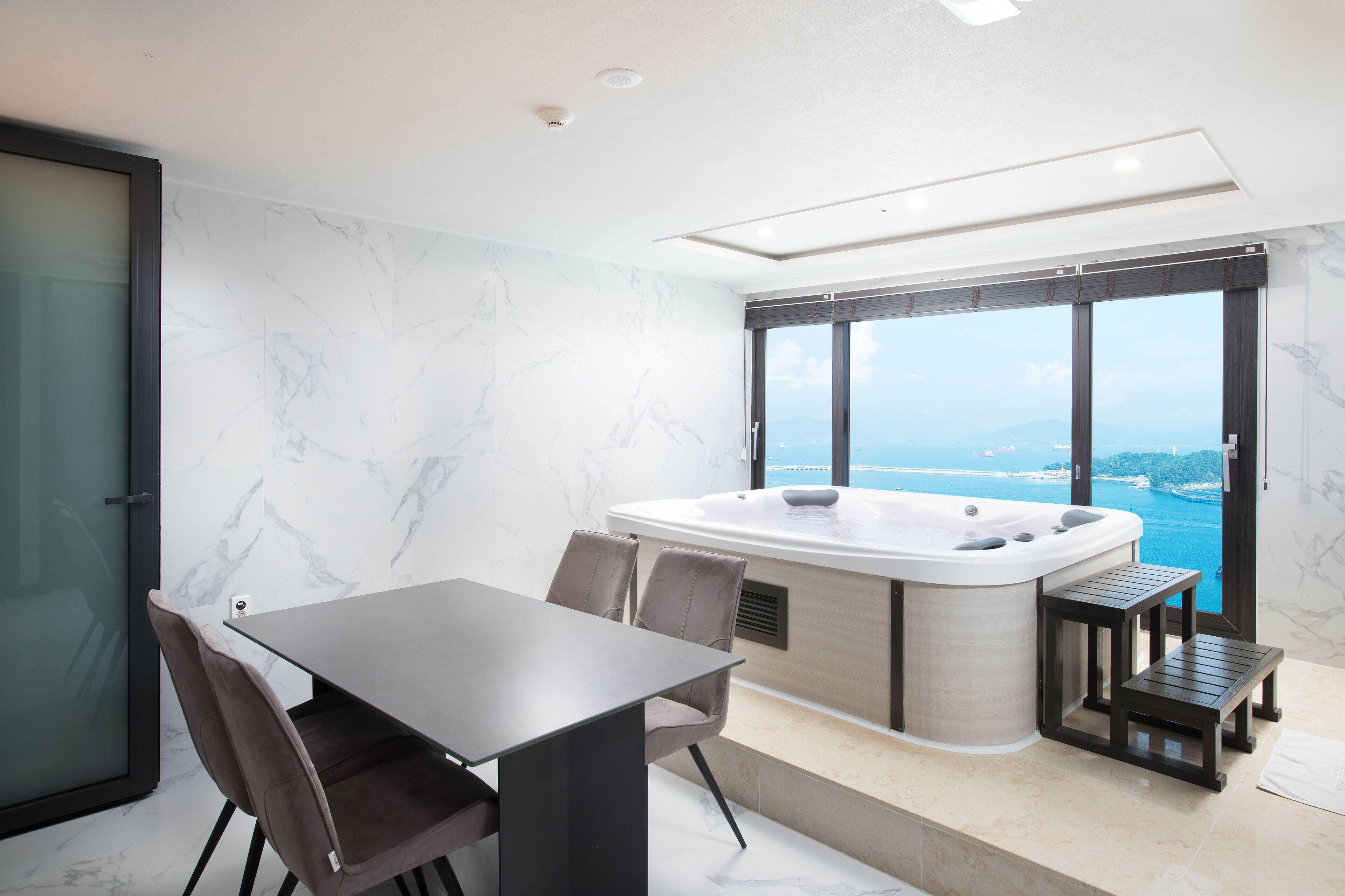 A modern room with a glass door, a dining table with chairs, a hot tub, and a view of the sea.