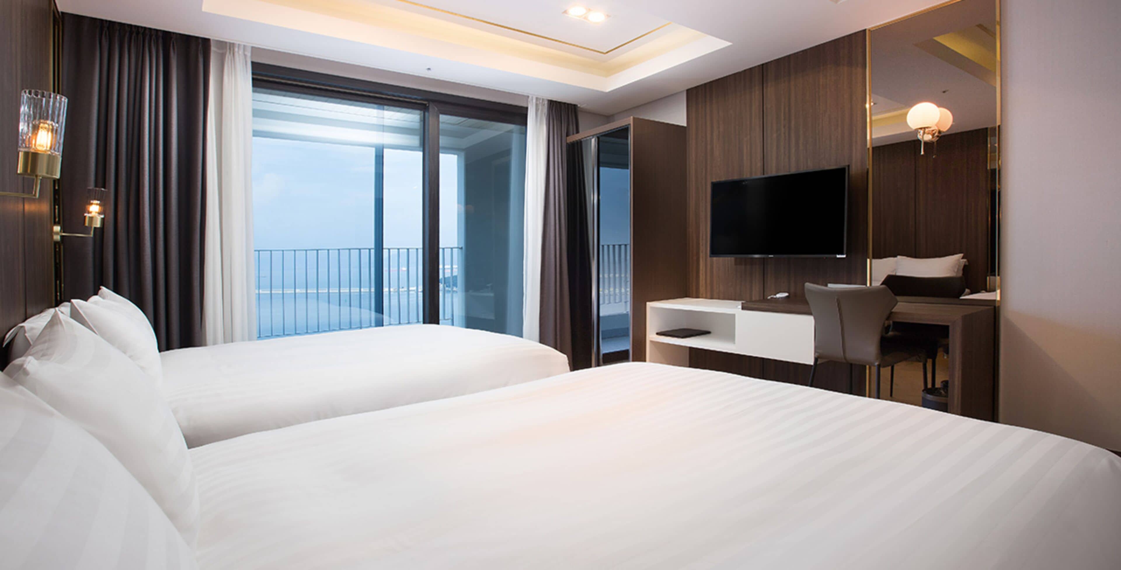 A hotel room with two beds, a desk, a chair, a TV, and a balcony with curtains.