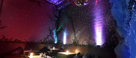 Un salón tenuemente iluminado con una bola de discoteca, iluminación ambiental verde y una pared de ladrillo con plantas.