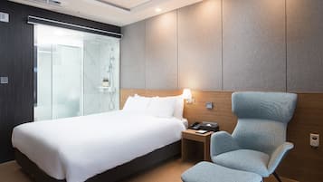 Habitación de hotel con cama, una silla, mesita de noche y cabina de ducha de vidrio.