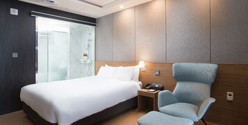 Habitación de hotel con cama, una silla, mesita de noche y cabina de ducha de vidrio.