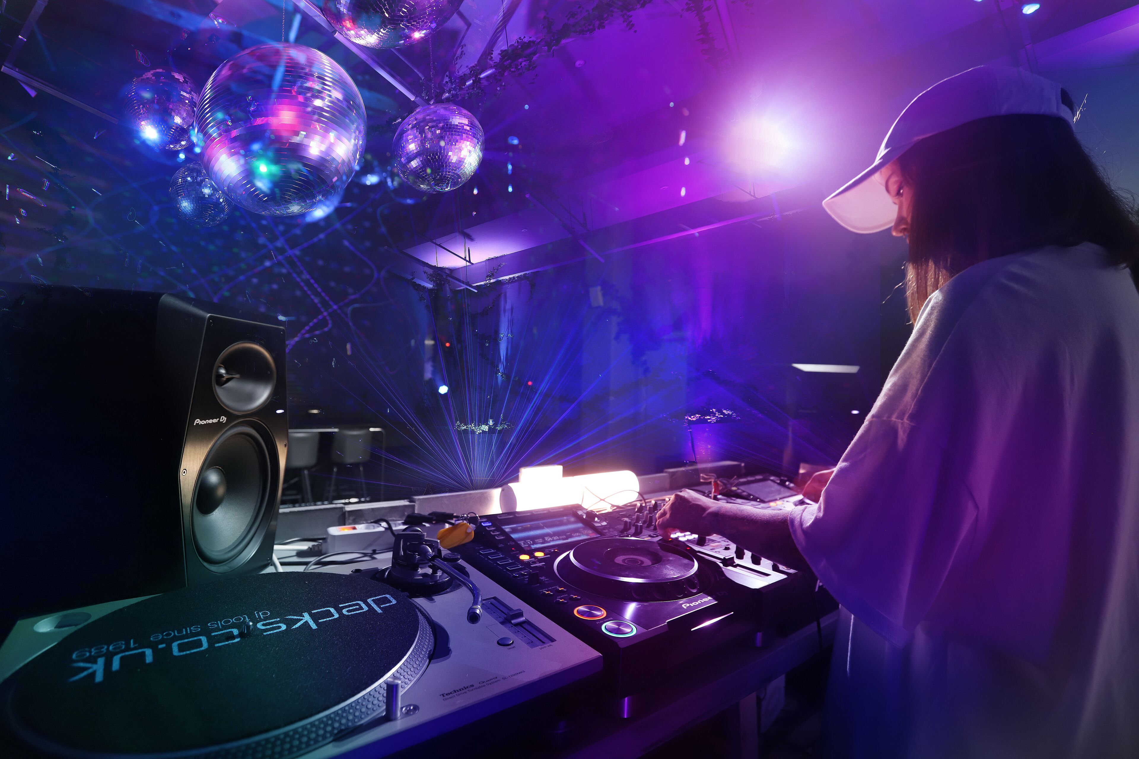 A DJ booth with a Pioneer DJ controller, a Pioneer DJ speaker, and a disco ball in a dimly lit room with purple lighting.