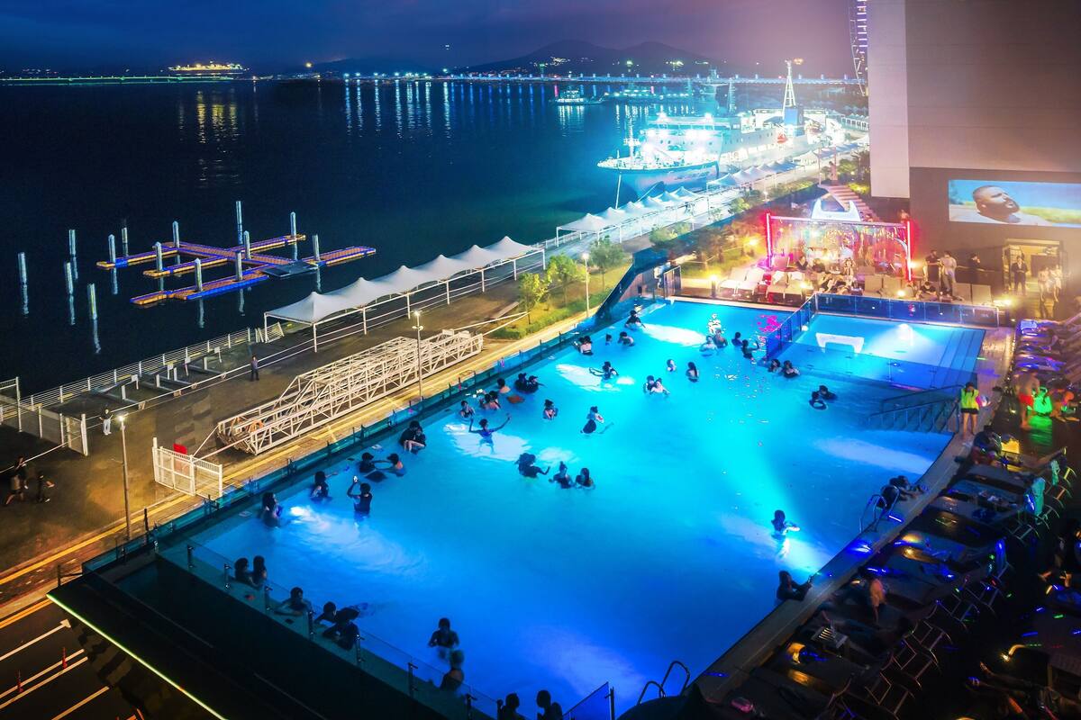 Een buitenzwembad met mensen die van het water genieten, omringd door een steiger en een podium voor evenementen.