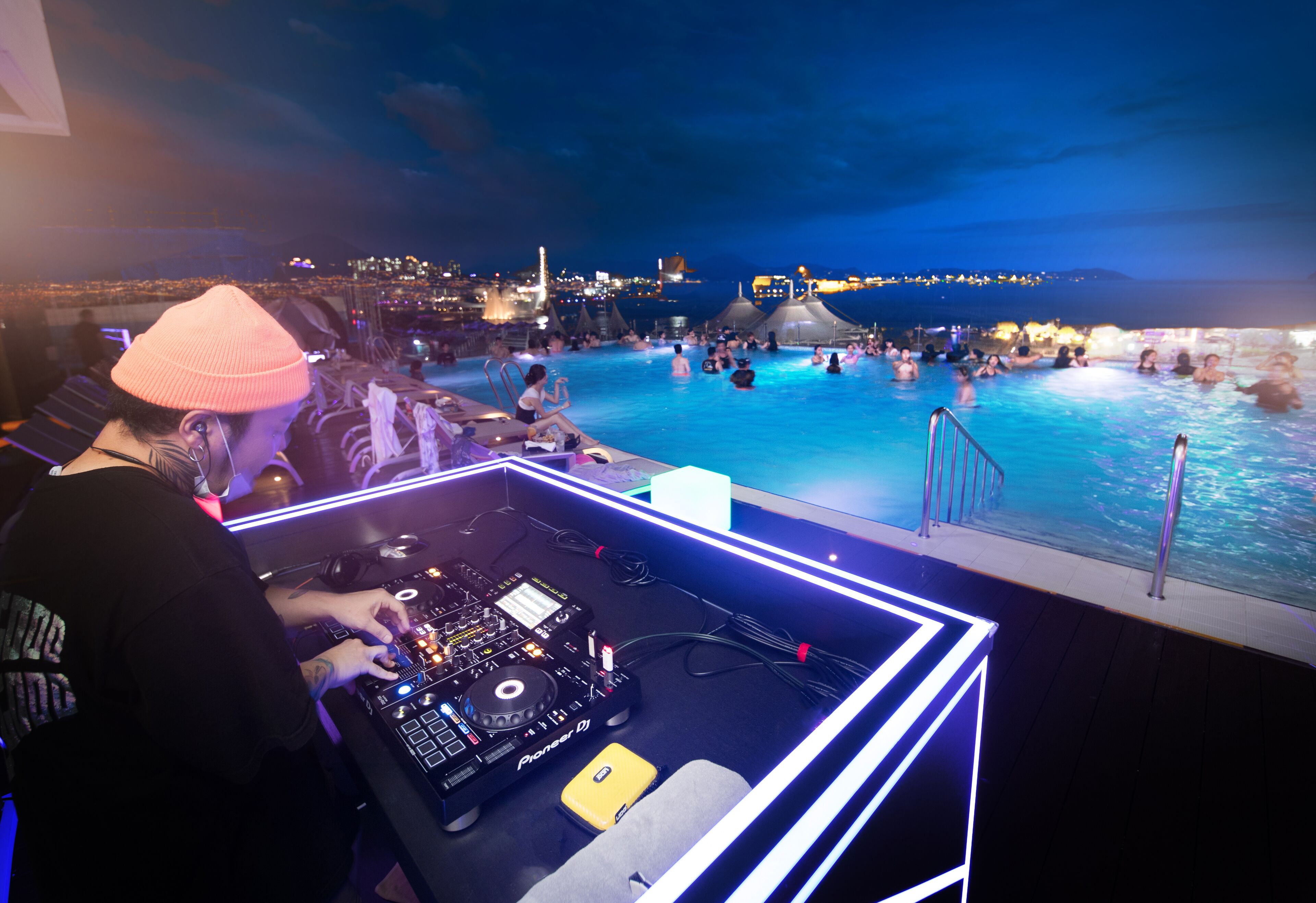 A DJ booth with a cityscape view, a pool area with people, and a sunset or sunrise sky.