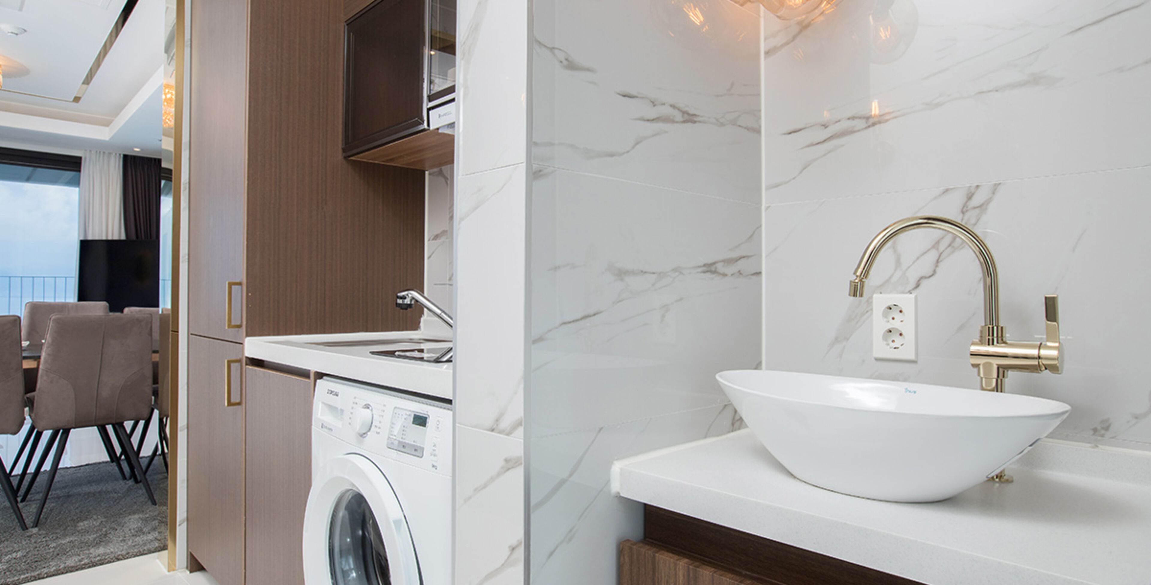 A modern bathroom with a white sink, gold faucet, and a washing machine.