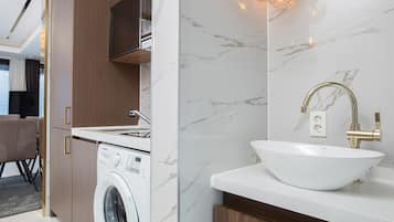 A modern bathroom with a white sink, gold faucet, and a washing machine.