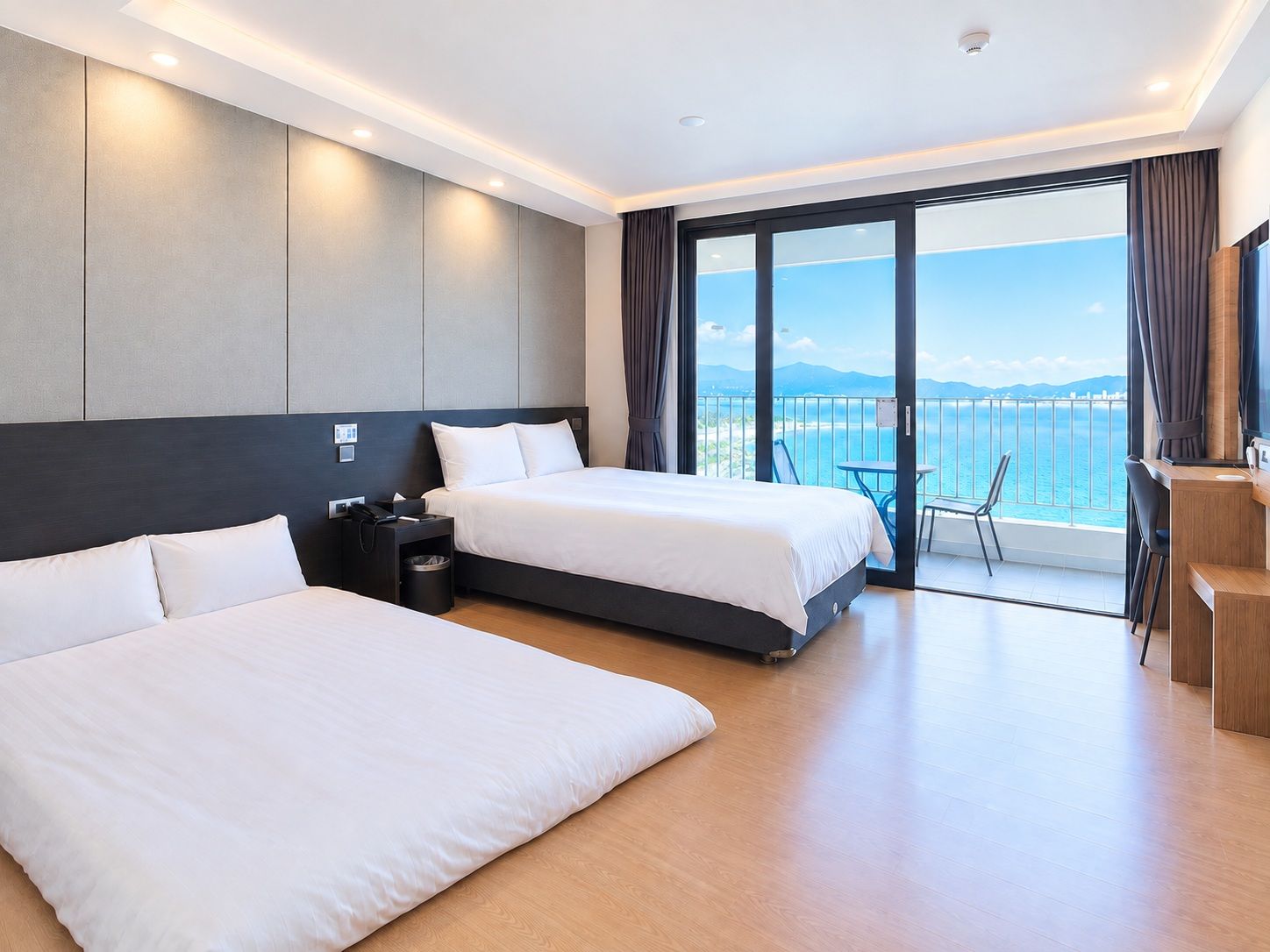 A hotel room with two beds, a desk, a chair, and a balcony with a view of the sea.