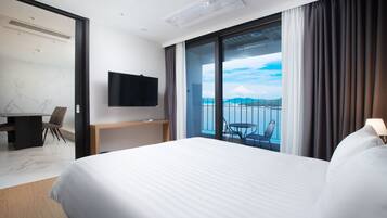 A hotel room with a large bed, a desk with a chair, a TV mounted on the wall, and a balcony with a view of the sea.