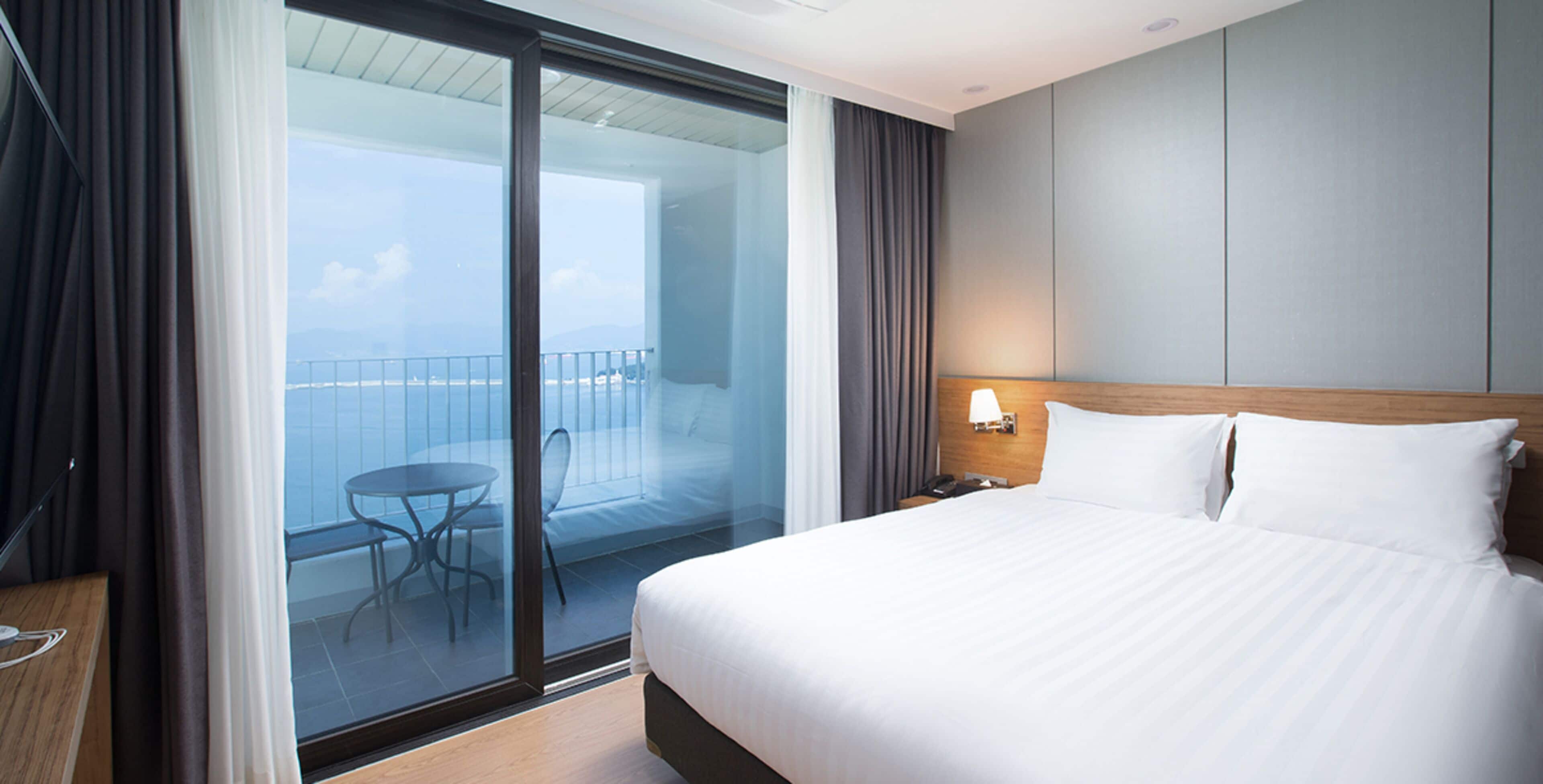Habitación de hotel con una cama grande, un balcón con mesa y sillas, y vista al mar.