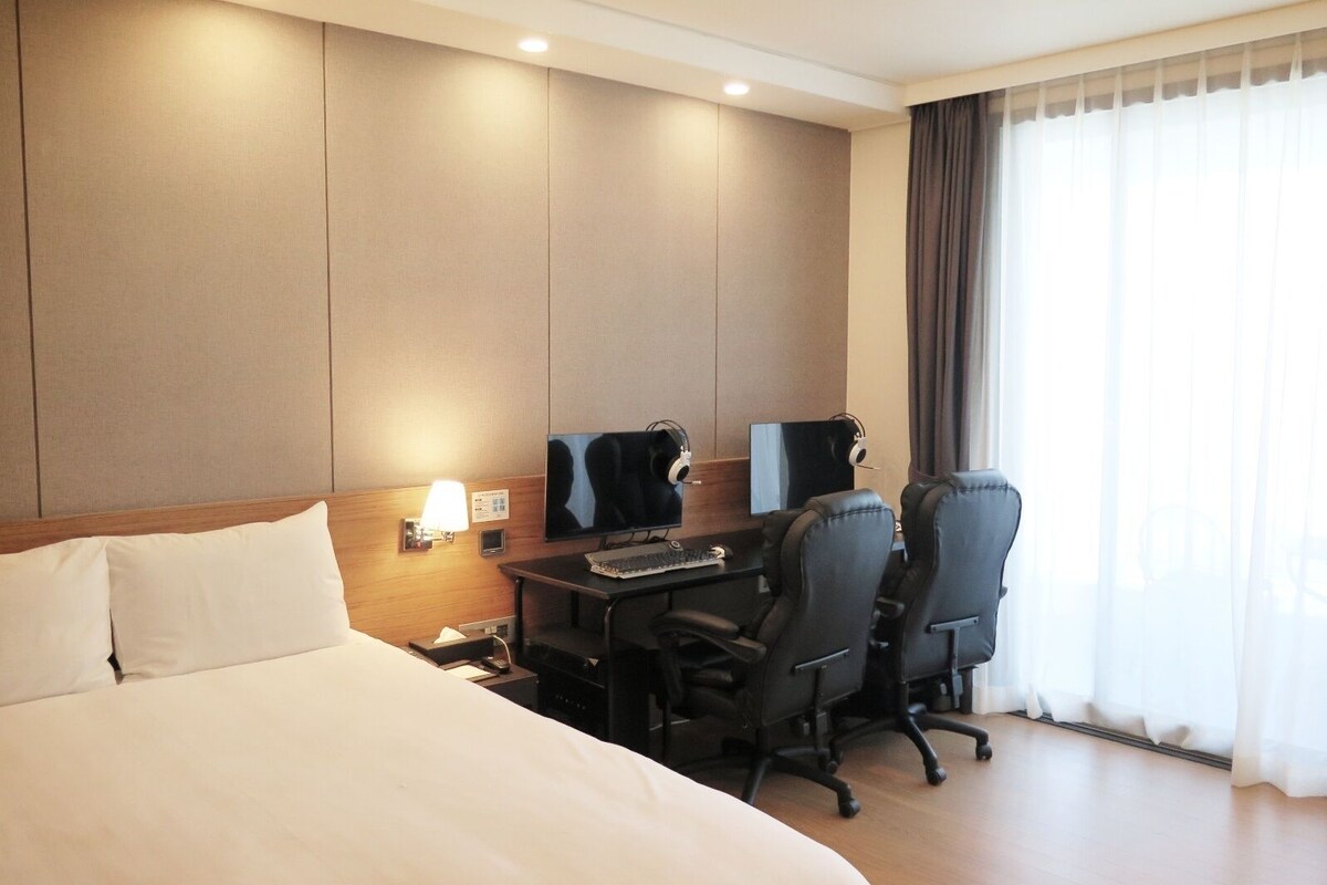 Habitación de hotel con cama, escritorio, dos sillas, televisor y ventana con cortinas.