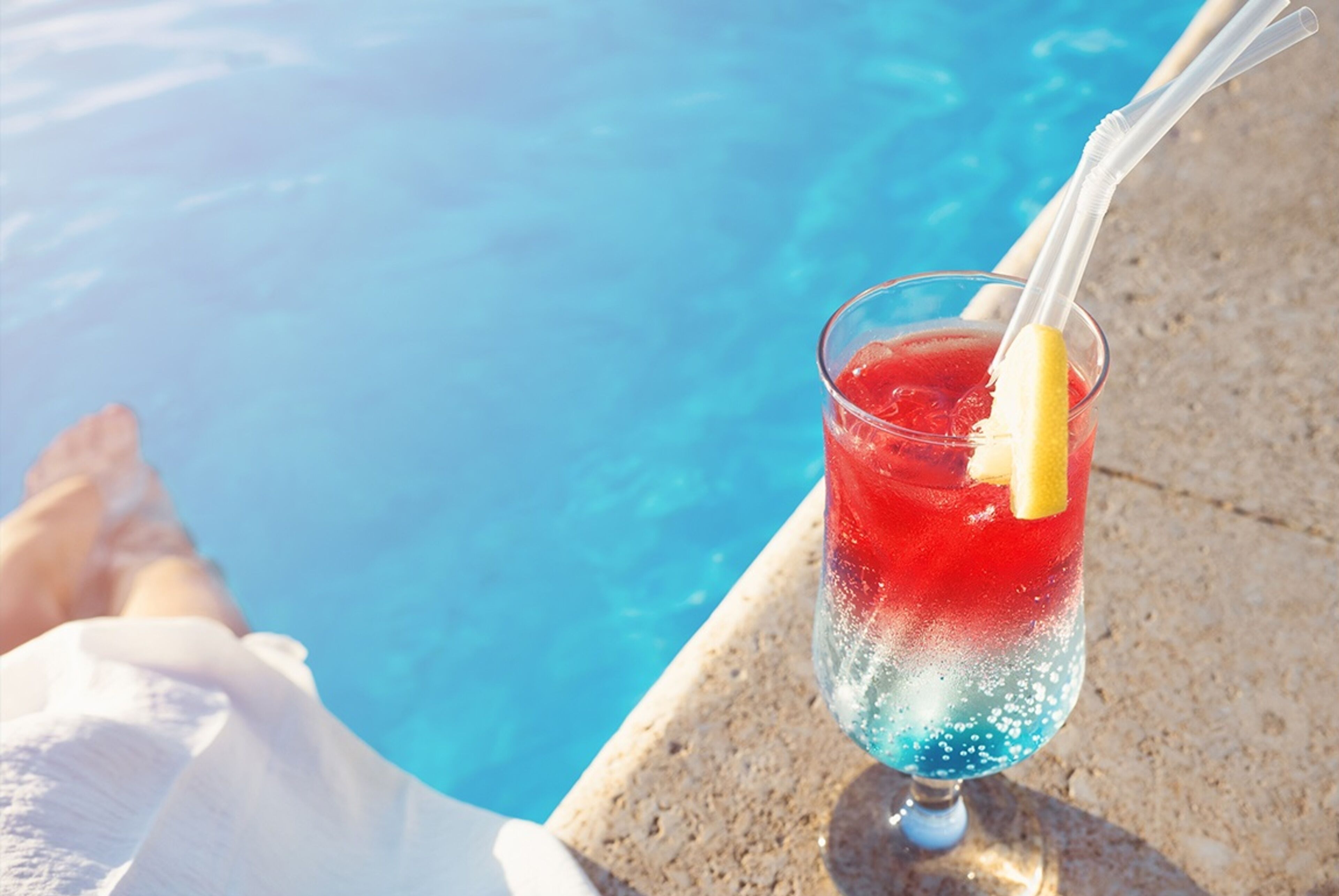 Un vaso de bebida roja y azul con una rodaja de limón y una pajita, sobre una barandilla junto a la piscina.