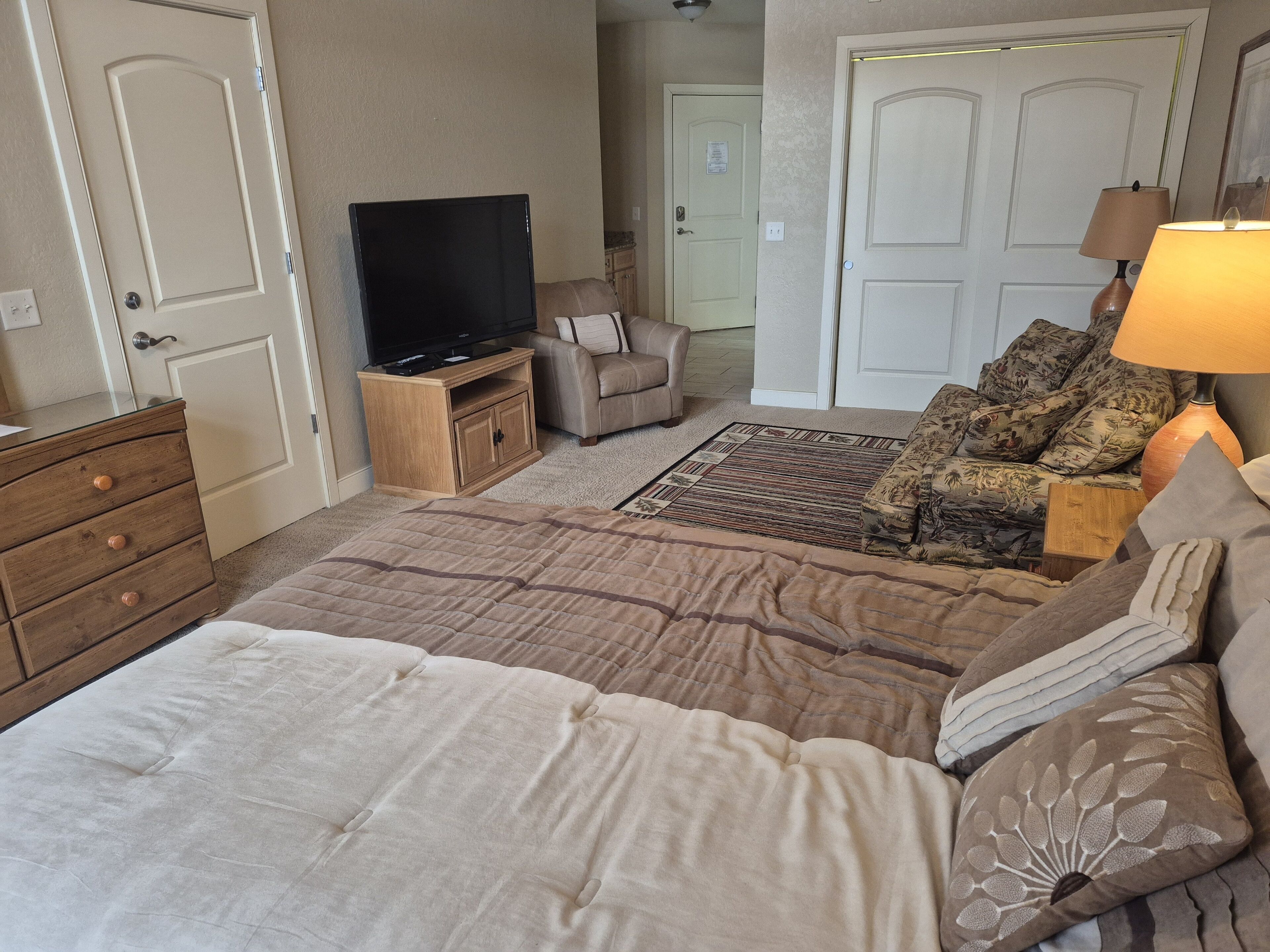 Een slaapkamer met een bed, een televisie op een houten standaard, een bank met decoratieve kussens, een lamp en een vloerbedekking.