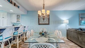 A dining area with a glass table, white chairs, and a chandelier. - Amelia by the Sea Oceanfront Condo with Access to Private Fishing Pier by RedAwning (Fernandina Beach)