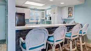 A kitchen with white cabinets, a black refrigerator, and a blue tiled backsplash. There are three wicker chairs with blue cushions at a bar area. - Amelia by the Sea Oceanfront Condo with Access to Private Fishing Pier by RedAwning (Fernandina Beach)