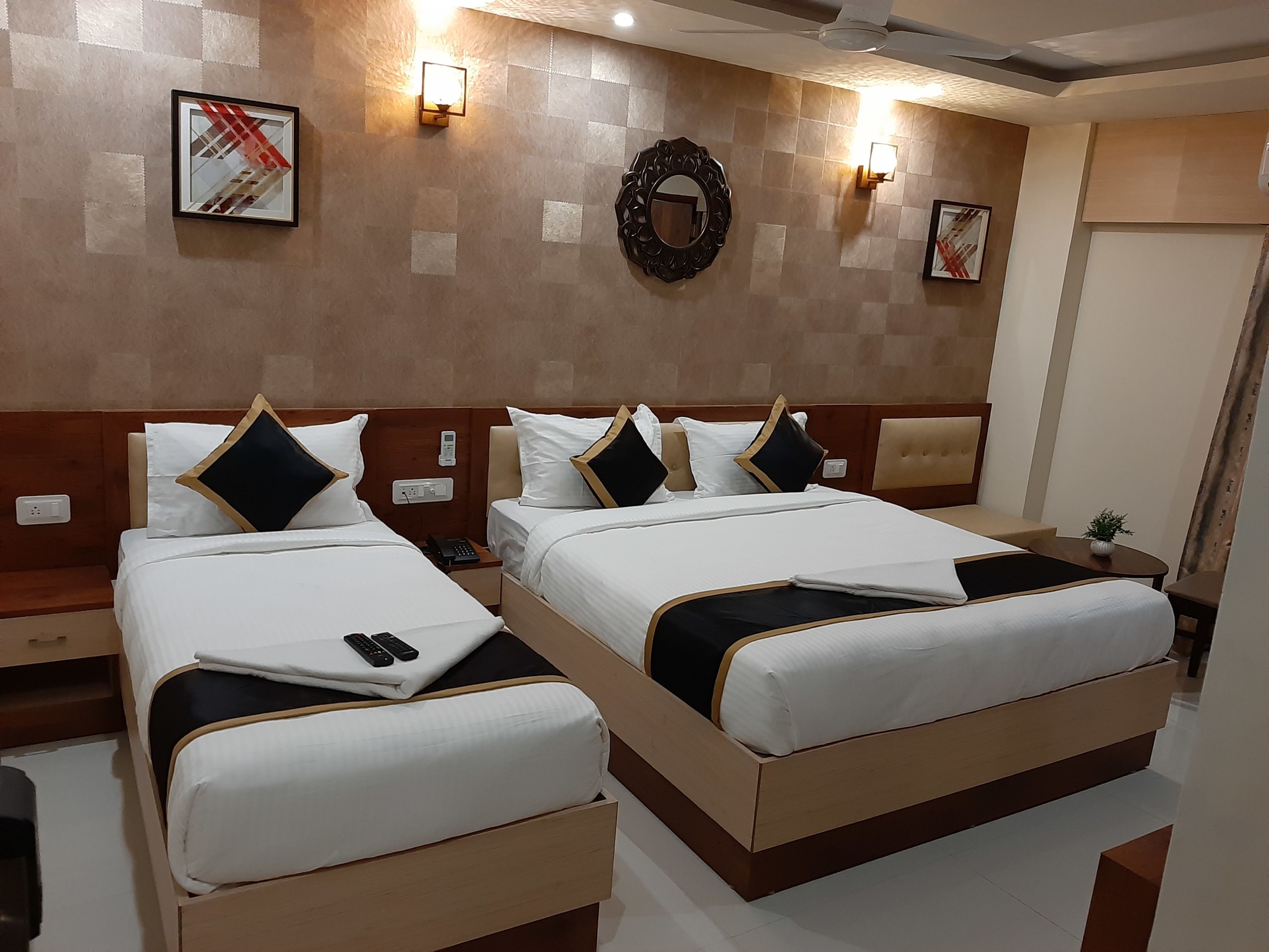 A hotel room with two beds, each with white bedding and black and gold pillows. There are wall-mounted lights, a mirror, and a framed picture on the wall.