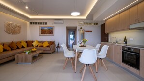 A modern living room with a sofa, coffee table, and dining area with chairs. There is a kitchen with wooden cabinets and a built-in oven. - Sirena Tolo Luxury Apartment (Nafplio)