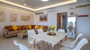 A modern living room with a sofa, dining table, and chairs. There are wall art pieces and an air conditioning unit. - Sirena Tolo Luxury Apartment (Nafplio)