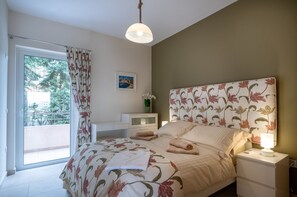 A bedroom with a bed, a nightstand, a bedside lamp, a window with curtains, a picture on the wall, and a view of greenery outside.