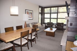 A modern living room with a wooden dining table, comfortable seating, a large window with blinds, and a television mounted on the wall. - Hayden Lodge  - Views Of The Slopes And Village 3 Bedroom Condo by RedAwning (Snowmass Village)