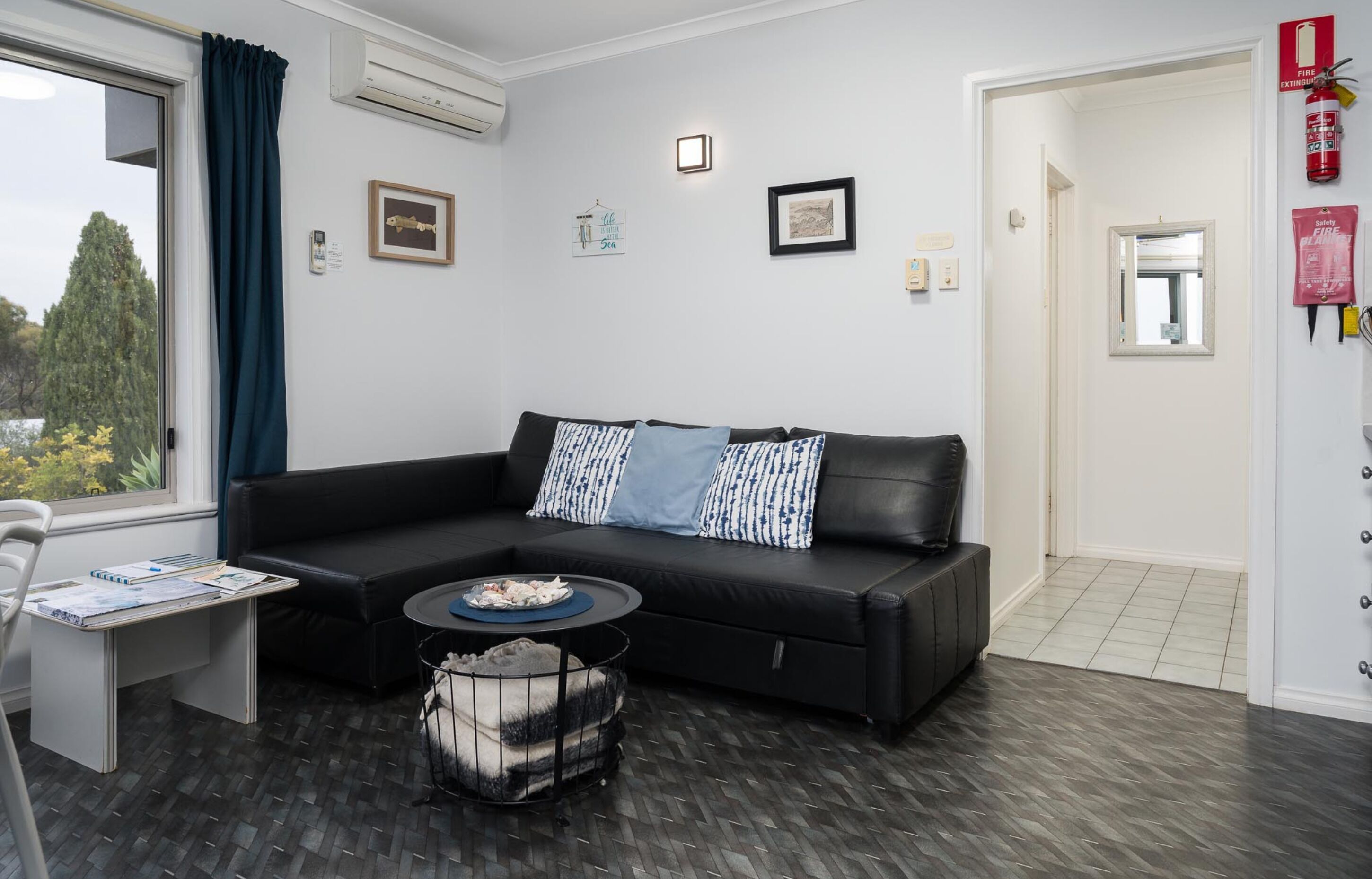 Ein modernes Wohnzimmer mit einem schwarzen Ecksofa, einem kleinen runden Couchtisch und einem Haustierbett. Es gibt ein Fenster mit Vorhängen, eine Tür mit Feuerlöscher und ein gerahmtes Bild an der Wand.