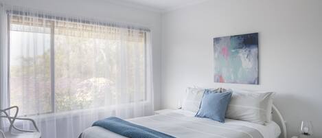 Una camera da letto con un letto, una sedia, un comodino, una ventola a soffitto e una finestra con tende leggere.