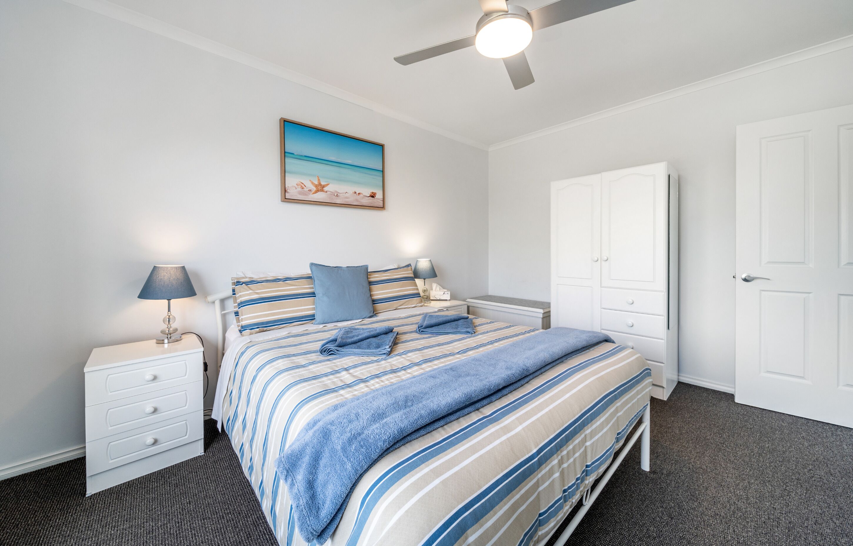 Een slaapkamer met een bed, nachtkastjes, een plafondventilator, een kledingkast en een schilderij aan de muur.