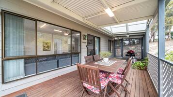 A covered patio with a wooden table and chairs, large windows, and a view of the outdoors.