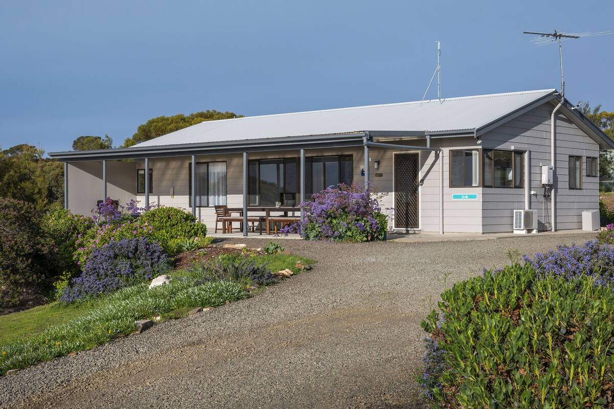 A modern building with a covered patio, a gravel driveway, and surrounding landscaped gardens.