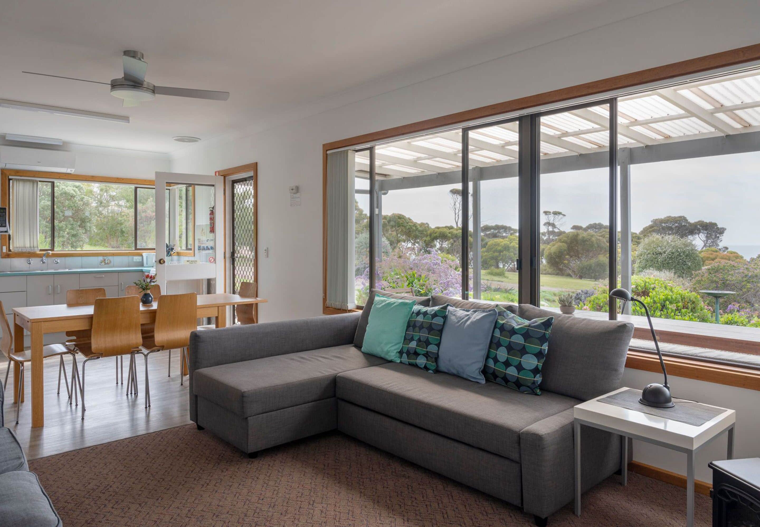 A modern living room with a sectional sofa, a dining table, and chairs. There is a large window with a view of greenery and trees.