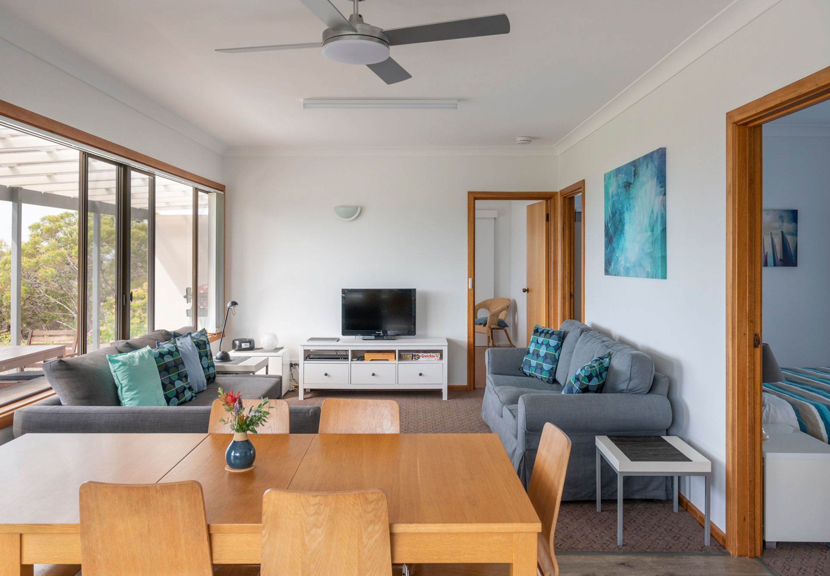 A modern living room with a dining area, large windows, a ceiling fan, and a television on a white cabinet.