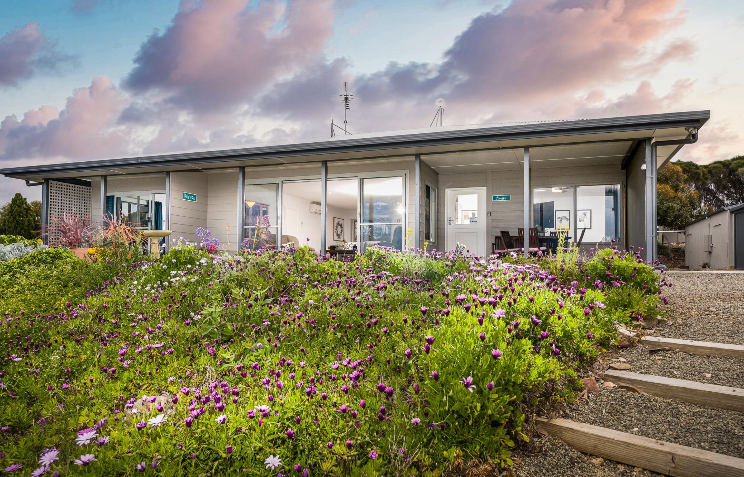 A modern building with a covered outdoor area, surrounded by a garden with purple flowers.