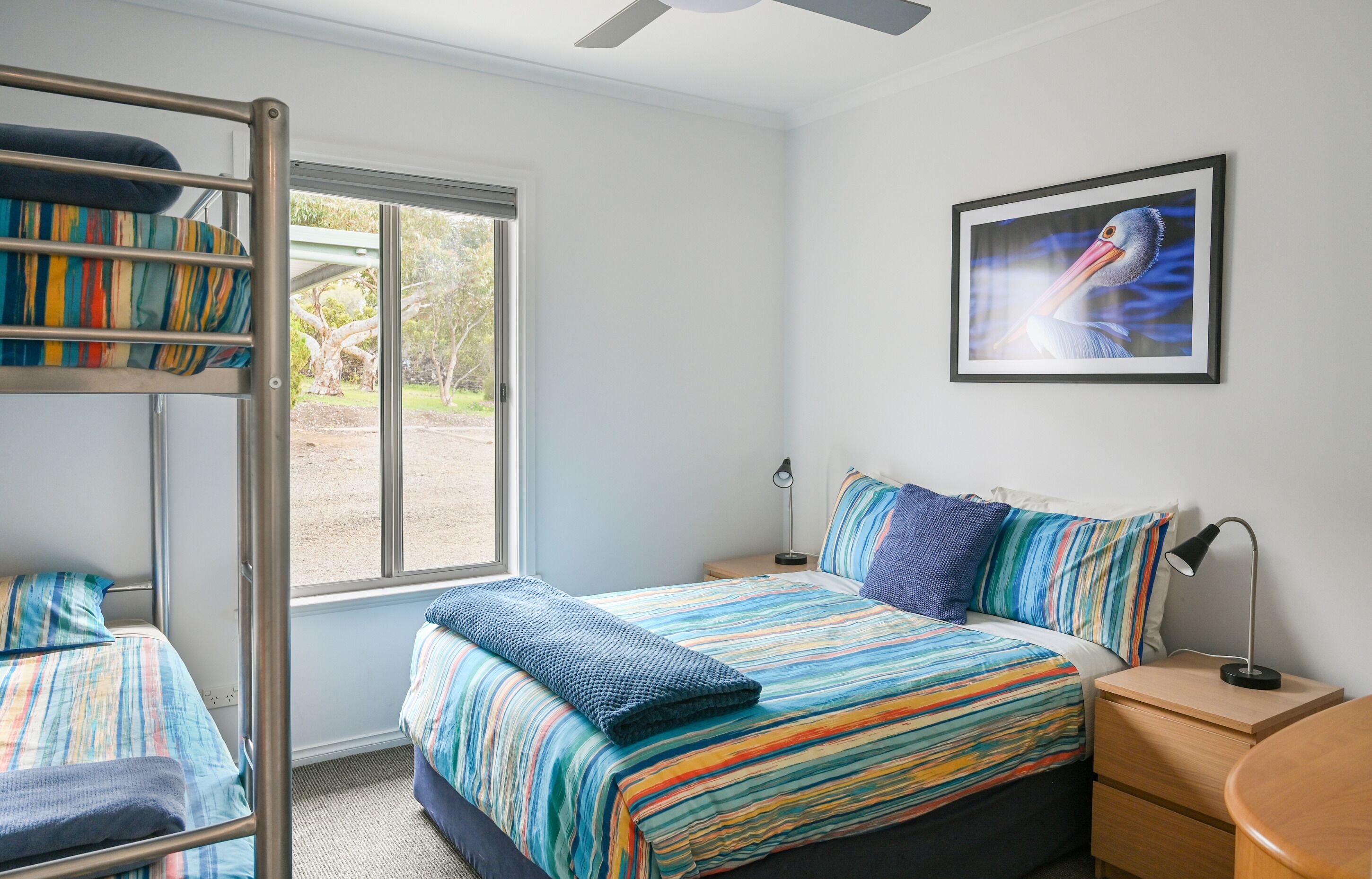 A bedroom with bunk beds, a bed with colorful bedding, a bedside table with a lamp, a framed picture on the wall, and a window with a view of greenery.