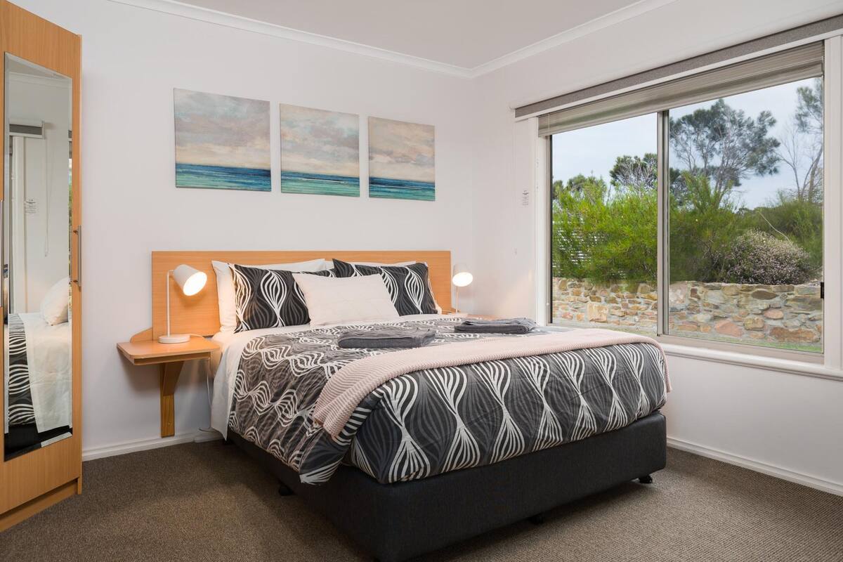 A bedroom with a bed, bedside lamps, a window with a view of greenery, and artwork on the wall.
