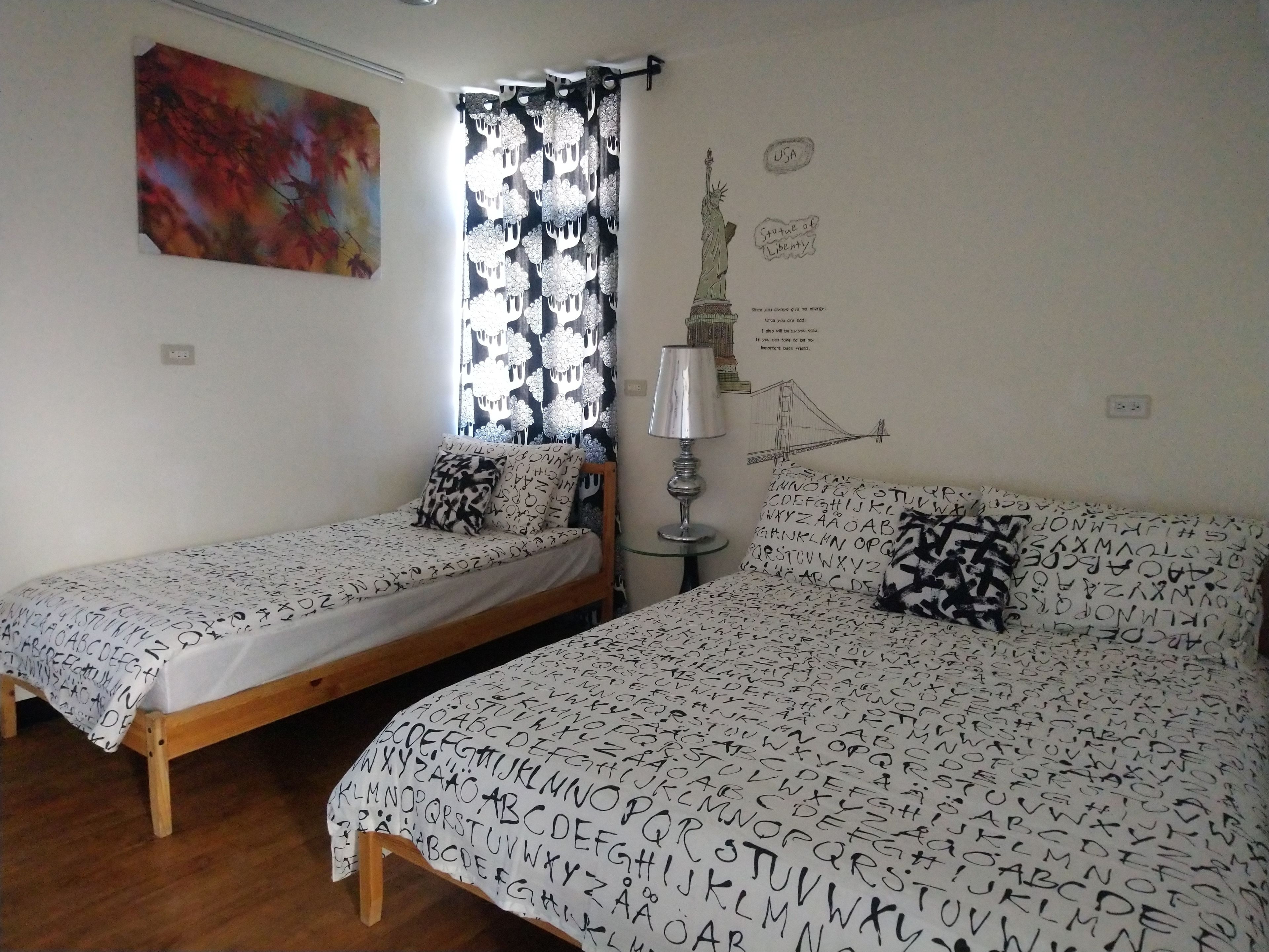 Ein Zimmer mit zwei Einzelbetten, einem Fenster mit gemusterten Vorhängen, einer Lampe und Wanddekorationen, darunter ein Gemälde und eine Zeichnung der Freiheitsstatue.