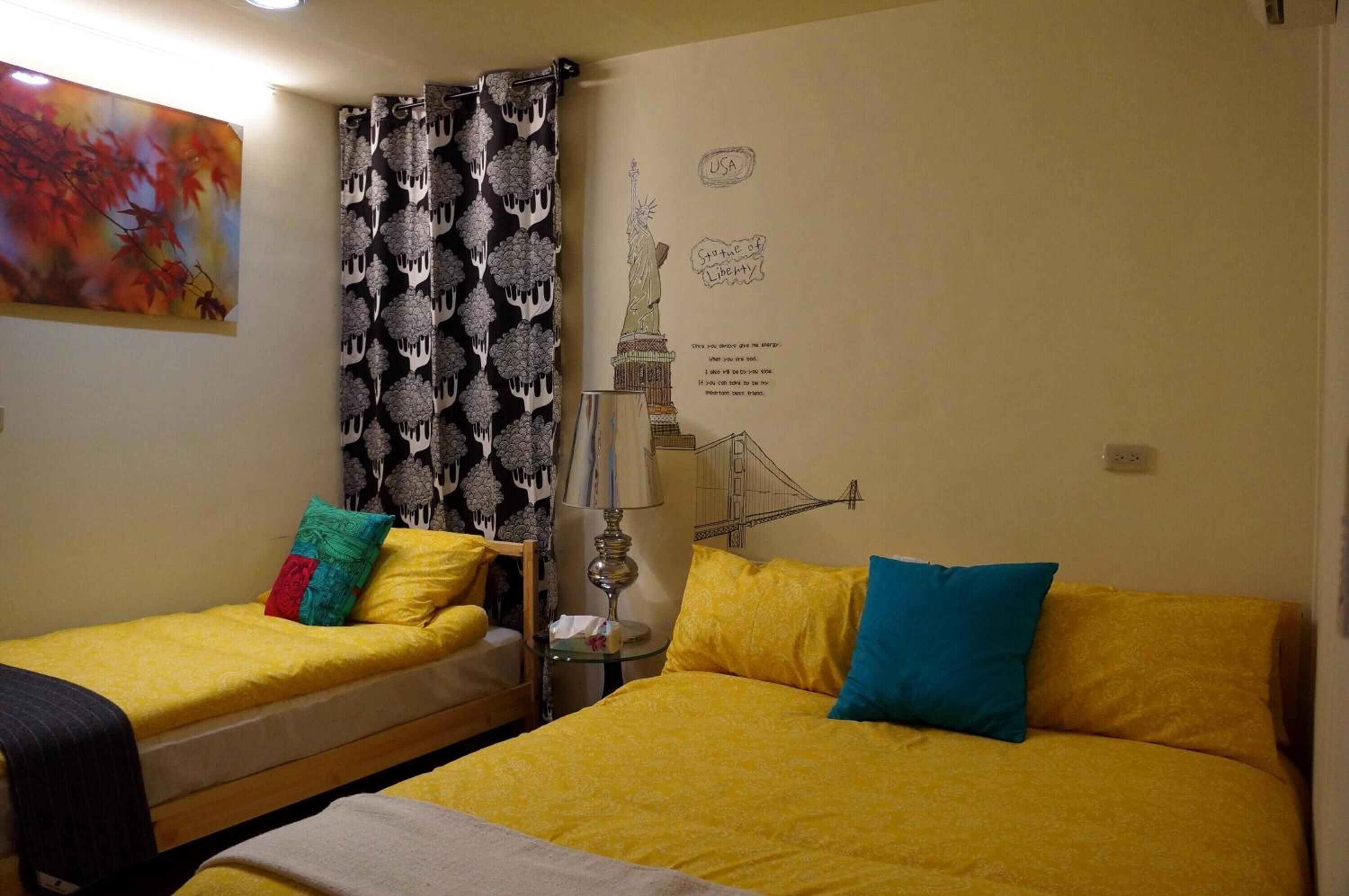 Ein Hotelzimmer mit zwei Betten, eines mit gelber Bettwäsche und das andere mit beiger Bettwäsche. An der Wand hängt ein Bild von Herbstlaub, ein schwarz-weiß gemustertes Vorhang und eine Wanddekoration der Freiheitsstatue.