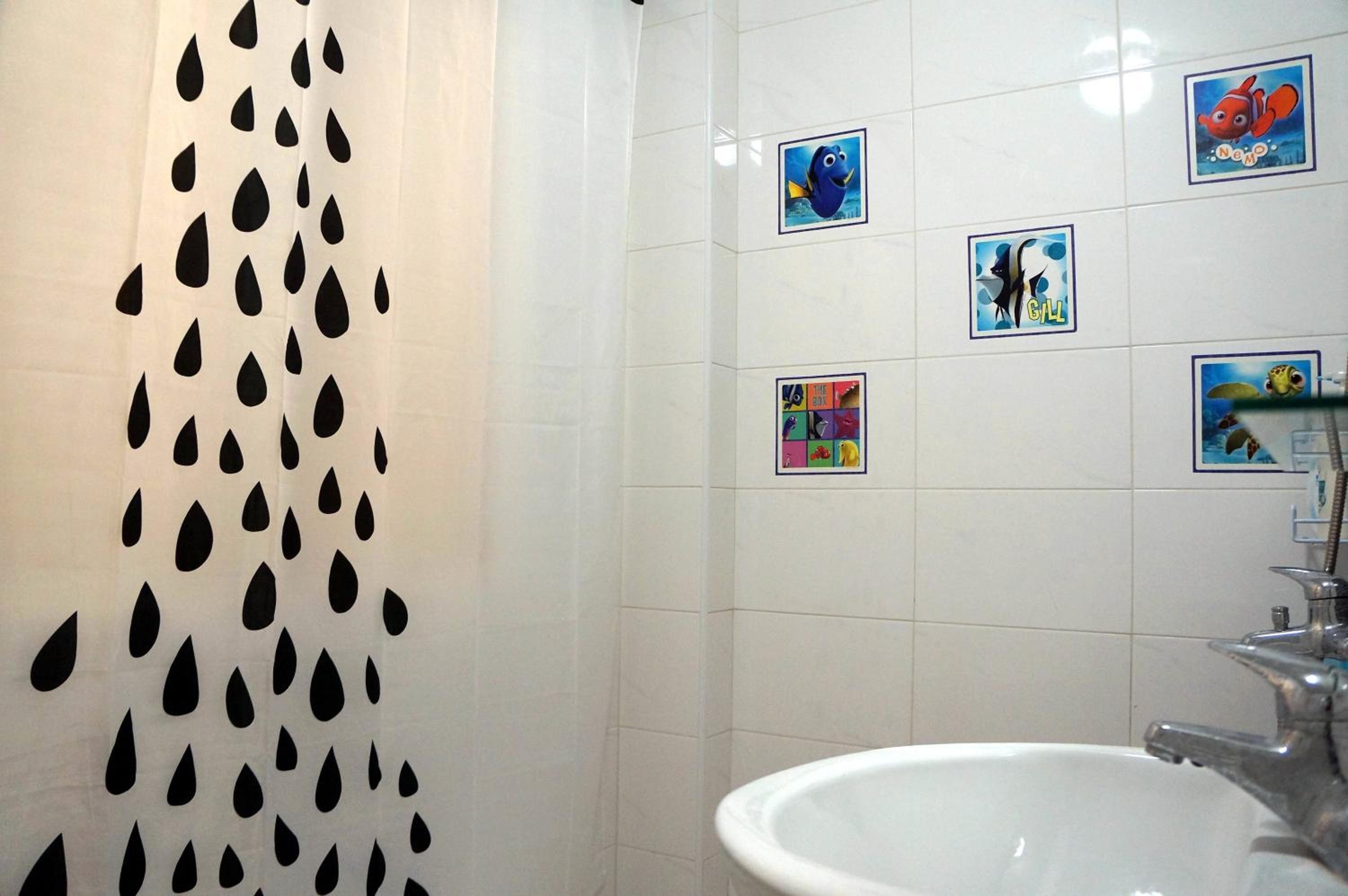 A bathroom with a white sink, a shower curtain with black teardrop patterns, and colorful wall stickers of various characters.