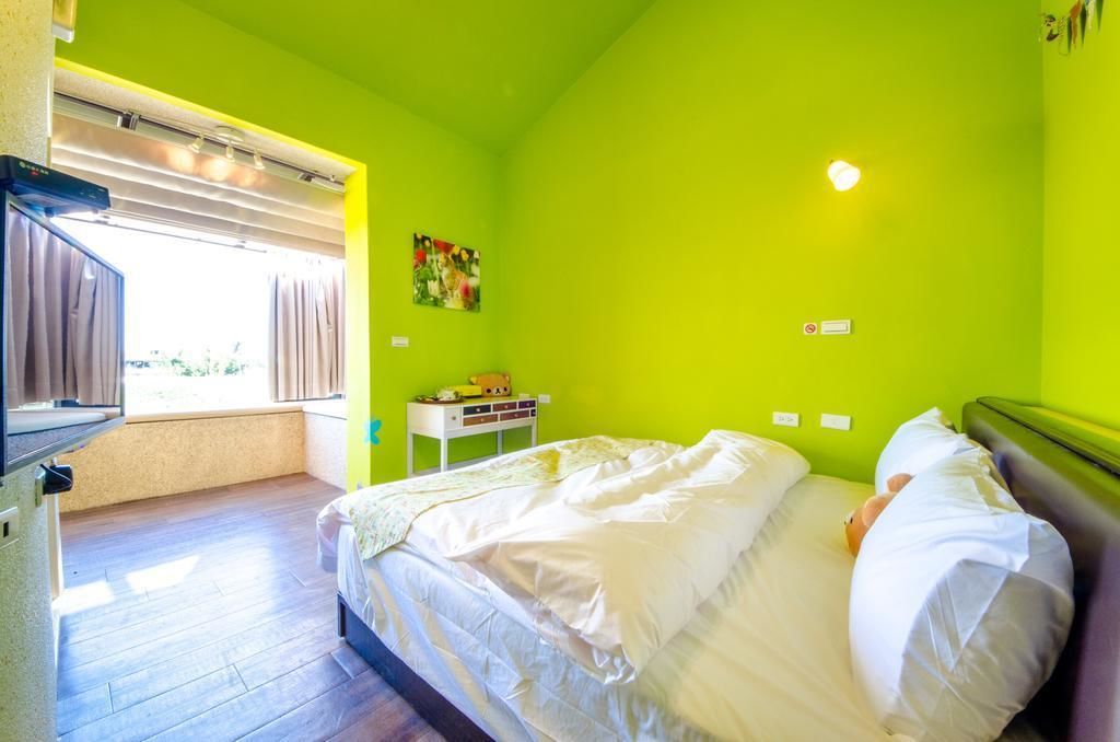 A room with a bed, a small table with a teddy bear, a window with curtains, and a green wall.