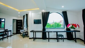 A modern living room with a large window, a flat-screen TV, a dining area, and a small table with a vase of flowers. - Hoianation Villas Hotel (Da Nang)